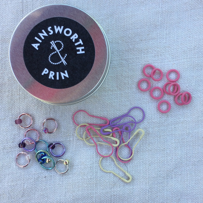 Three types of stitch marker, small pink rubber rings, pastel pear shaped pins and pastel rings with coordinating beads plus the aluminium tin.