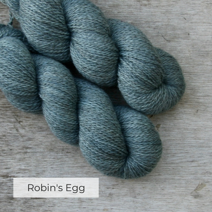 Two skeins of wool on a wood background. The yarn is a light blue green and the brown of the original wool shows through