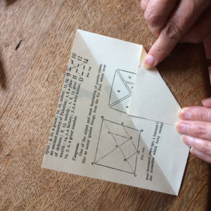 A pair of hands folding one corner of a square of paper up to the halfway line. The background is a wooden table