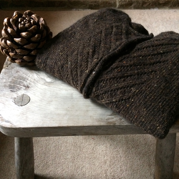 A brown tweedy cardigan folded neatly on a wooden stool with a large pinecone