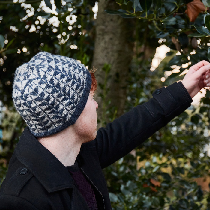 A man standing in front of a holly bush in a dark blue peacoat and wearing a blue and light grey beanie with an arrow design