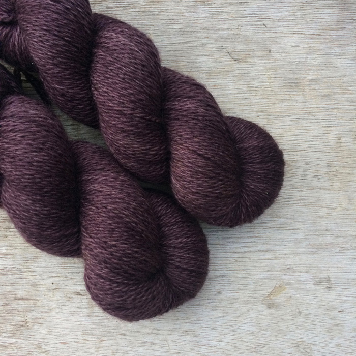 Two skeins of wool on a wood background. The yarn is the colour of a bottle of Damson Gin, purple but with brown too