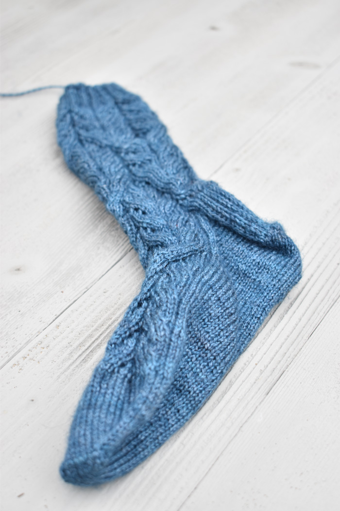 An unblocked blue hand knit lace sock on a white wood background