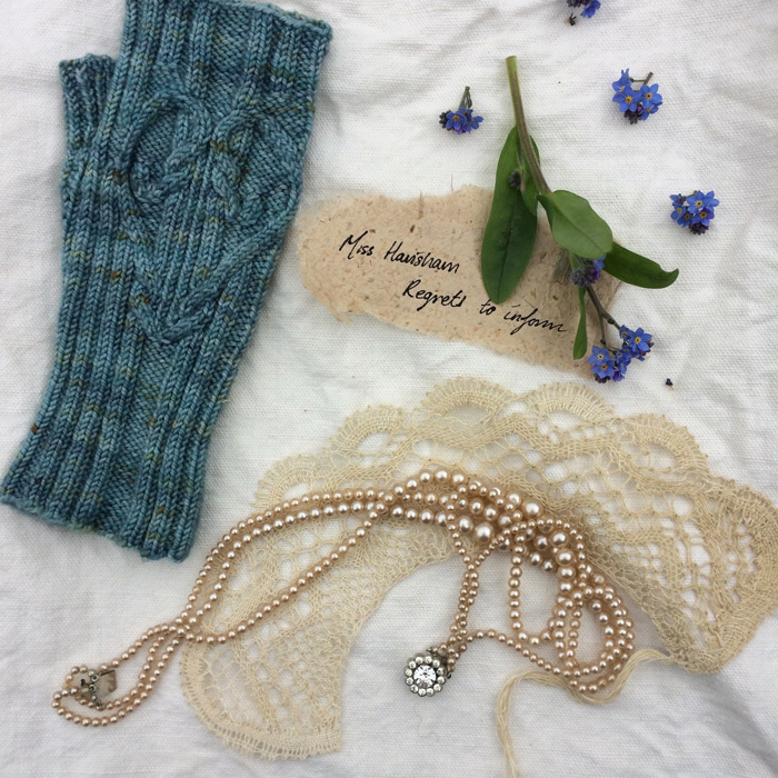 A single blue mitten with cabled heart motif , forget-me-not flowers, lace, pearls and Miss Havisham regrets card on white