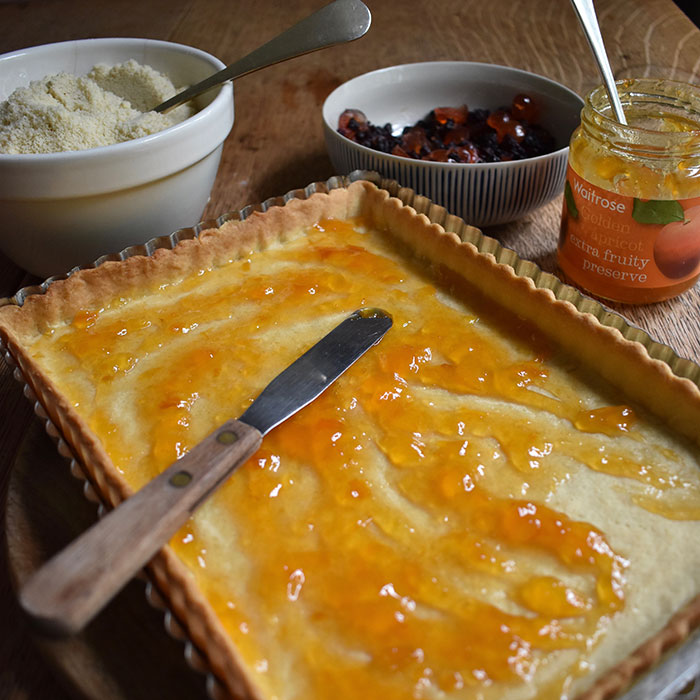 Pastry case spread with apricot jam with bowls and jam jar in background