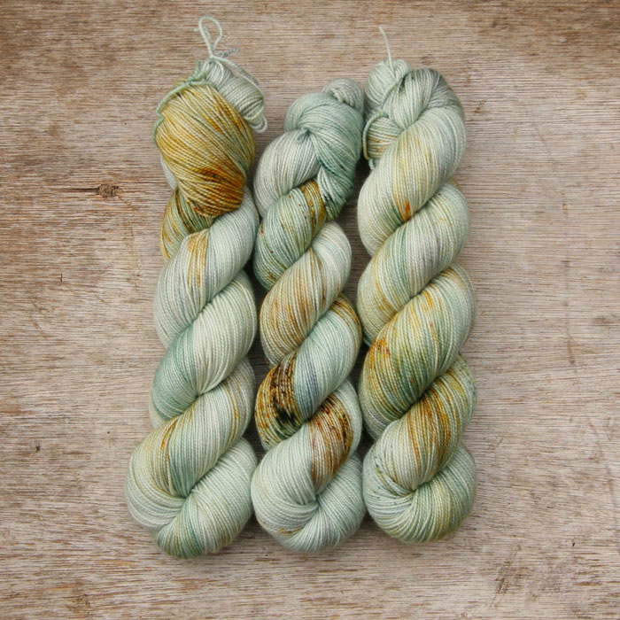Three skeins of sock yarn dyed in shades of mint, pearl grey and blue with speckles of dark green, rust and brown