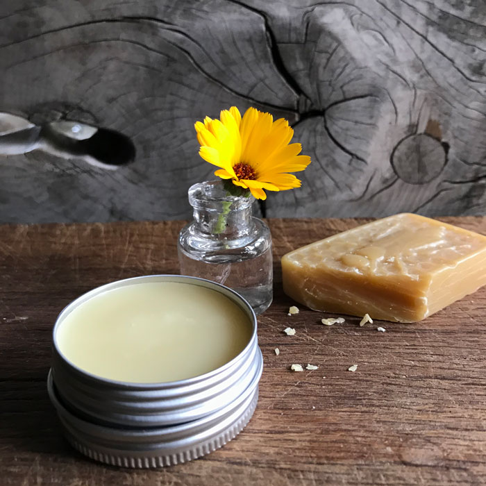 An open aluminium tin of a pale cream ointment a wooden block with a block of beeswax and a marigold in the background