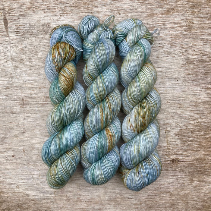 skeins of yarn dyed in shades of pale blue, green and grey with speckles of gold and deep green