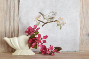 A cream vase with a branch of pink apple blossom that frames an embroidery of a bird on a branch