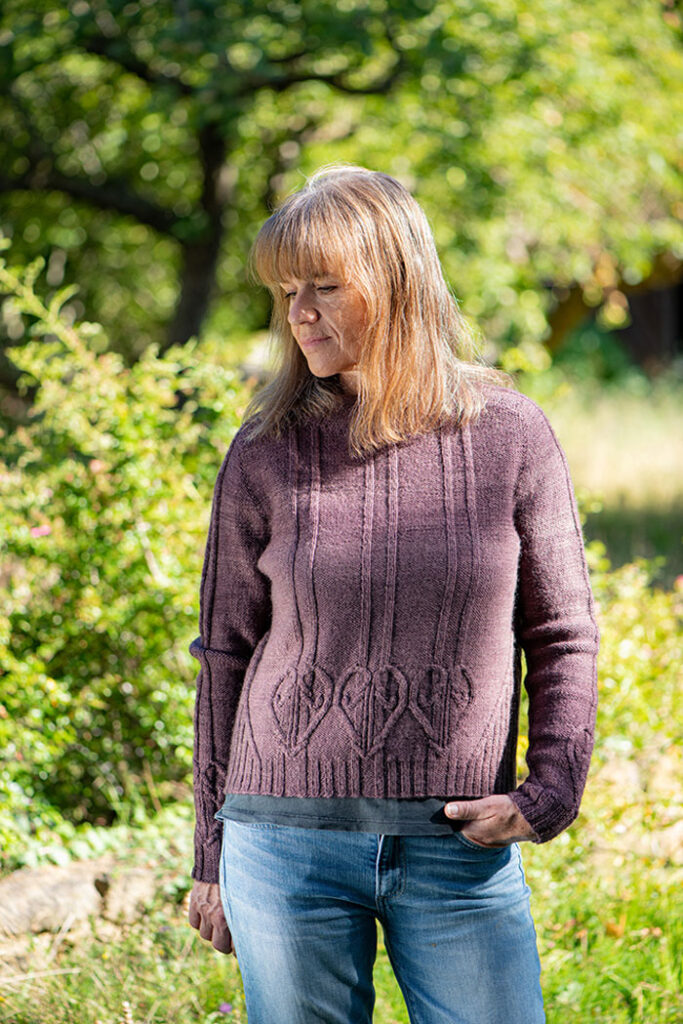 A woman standing in the shade of a tree wearing a long sleeved jumper with heart shaped cables at the hem