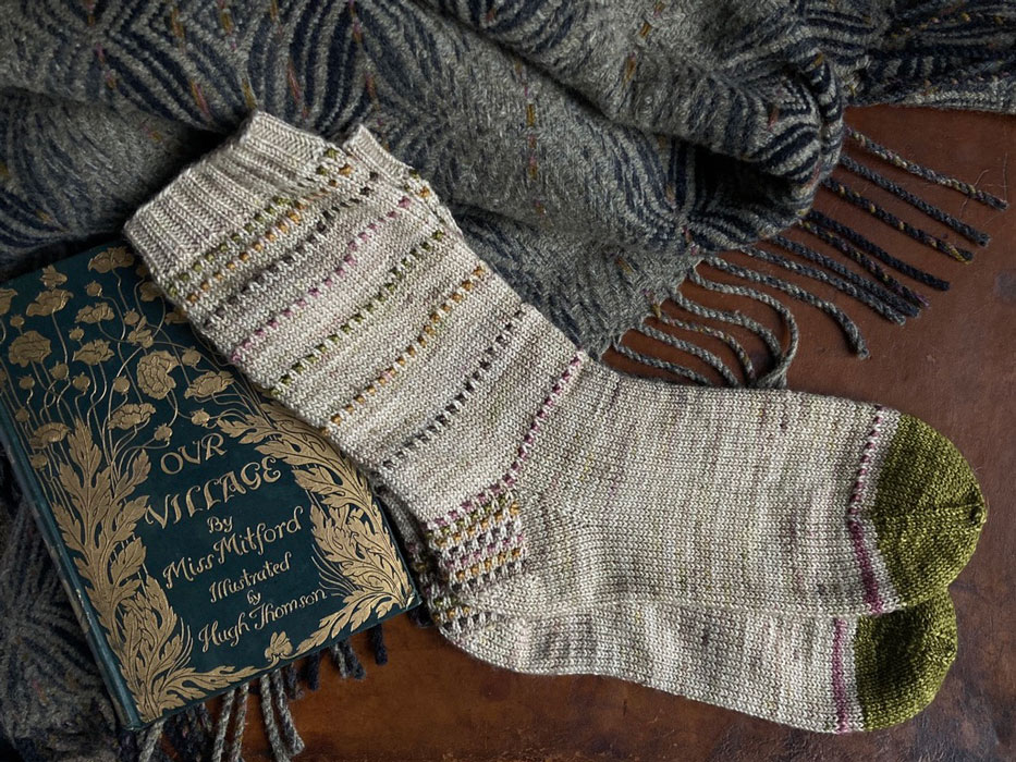 A pair of hand knit socks laying on an old leather suitcase with a throw and old book