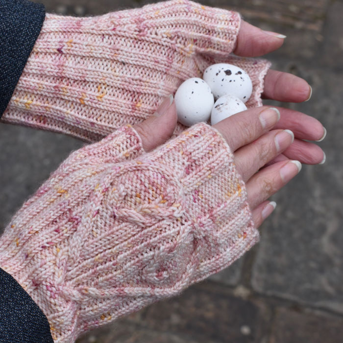 A pair of hands wearing pink fingerless gloves with a cabled heart motif and holding some small eggs