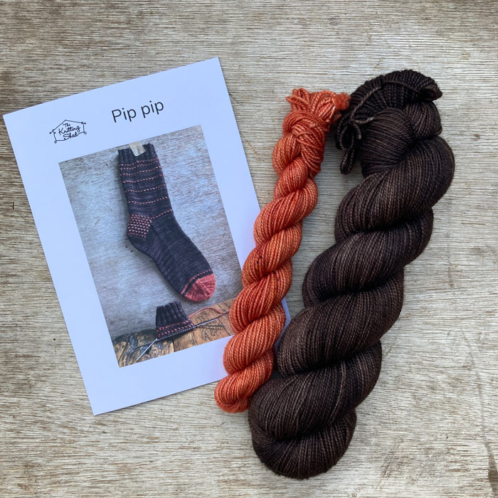A wooden background with a copy of a printed pattern plus a sock set to knit Pip Pip socks