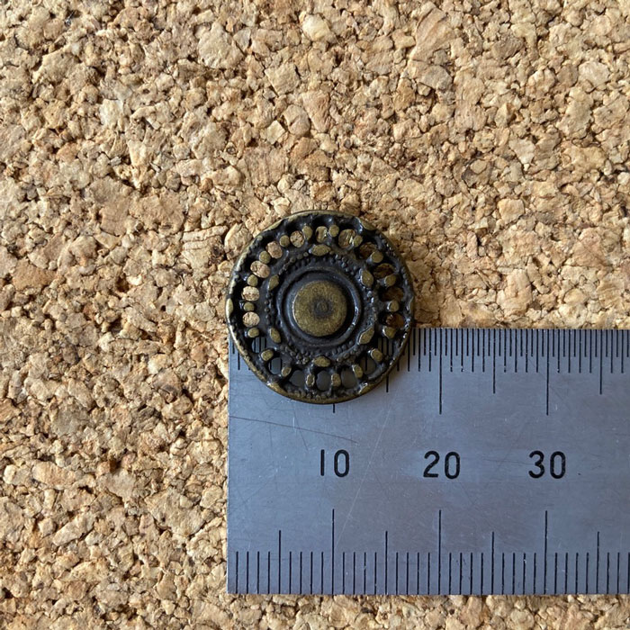 A pierced metal shank button against a ruler