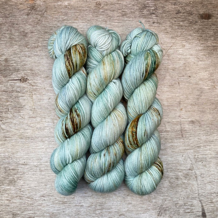 Three skeins of yarn dyed in shades of mint, pearl grey and blue with speckles of dark green, rust and brown