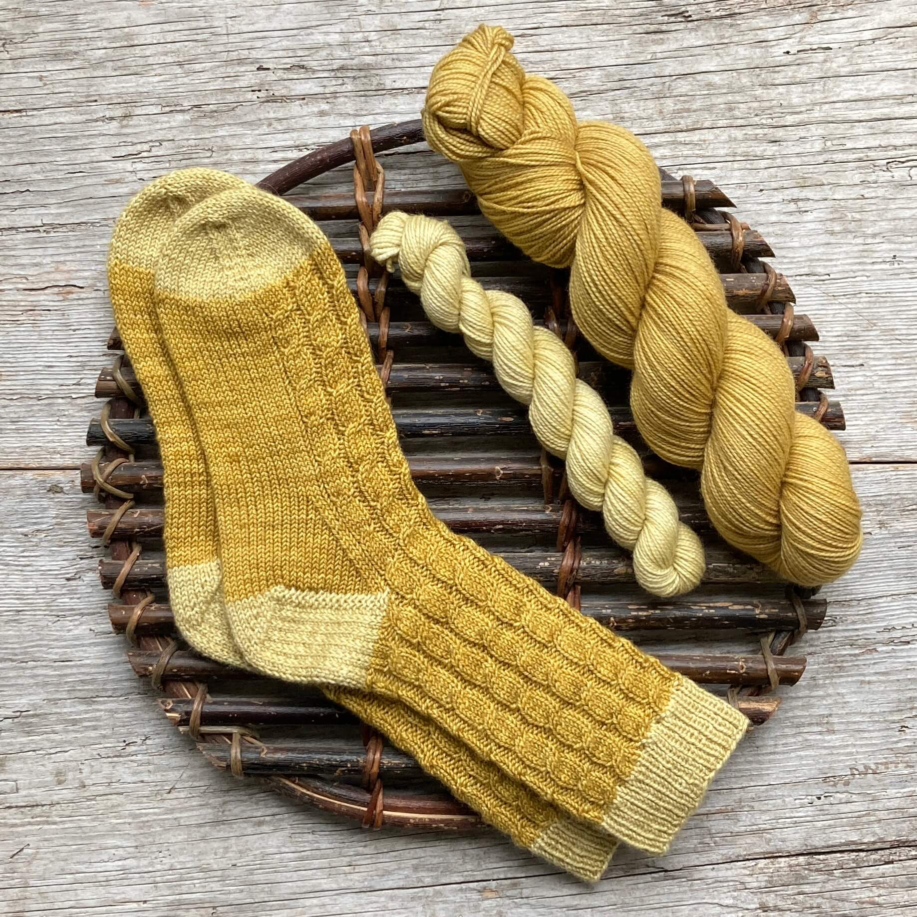 A pair of hand knit cabled socks with contrasting cuff, heels and toes and the yarn used to make them on a willow platter