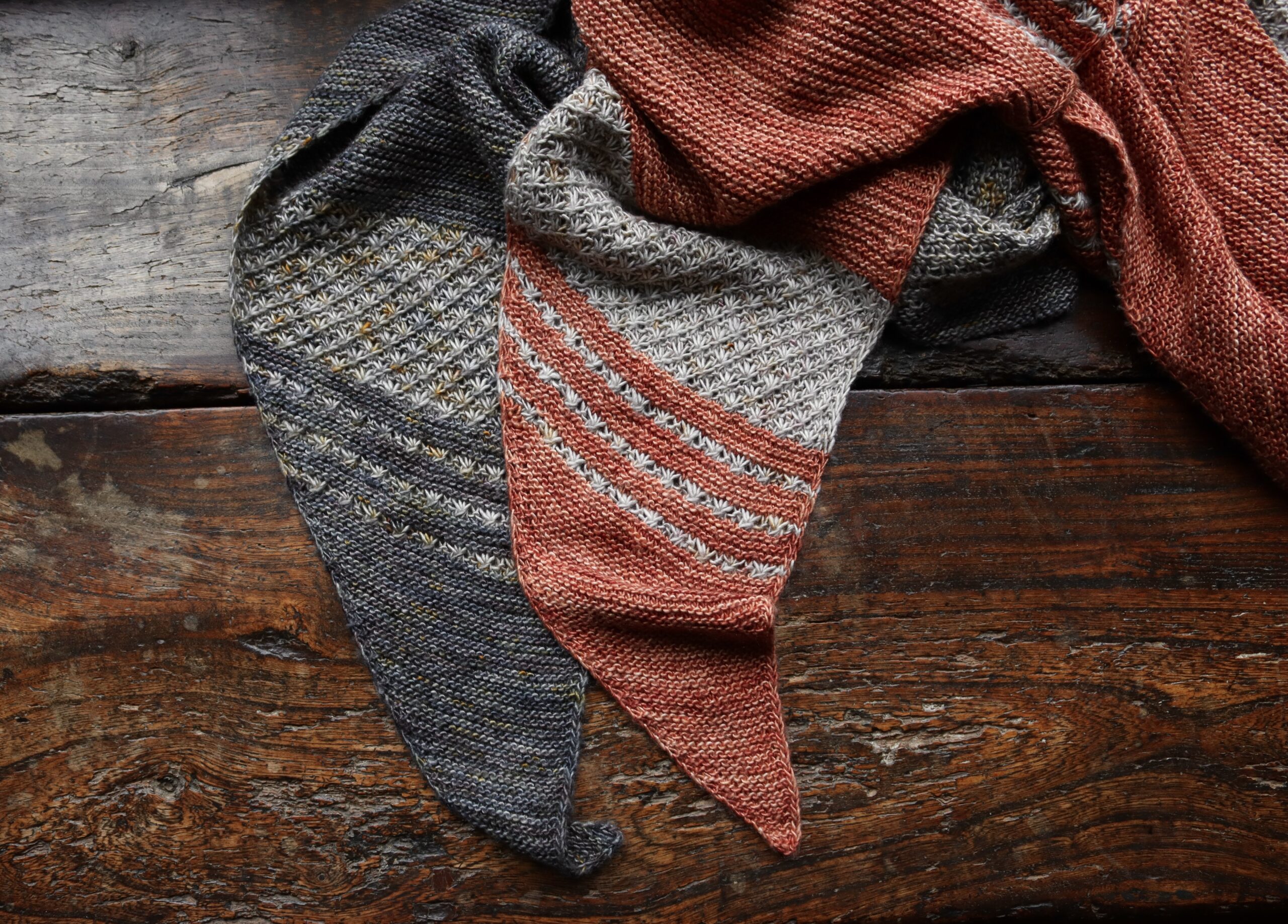 Two knitted shawls on a dark wood background, one in grey and the other terracotta