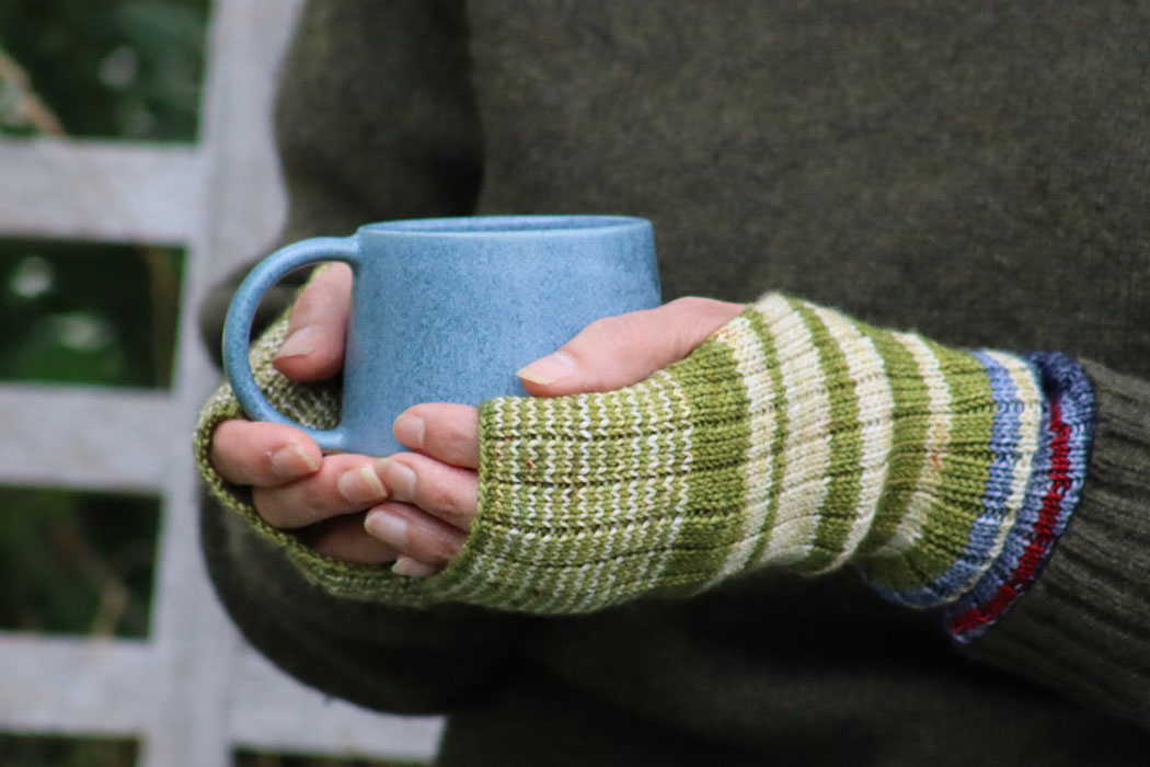 A close up of a pair of women's hands wearing a pair of stripy hand warmers and holding a pale blue mug