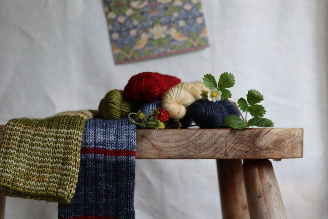 Five different coloured mini skeins and a pair of ribbed wrist warmers on a wooden stool decorated with strawberry fruits, leaves and flowers. Behind is the card that inspired them