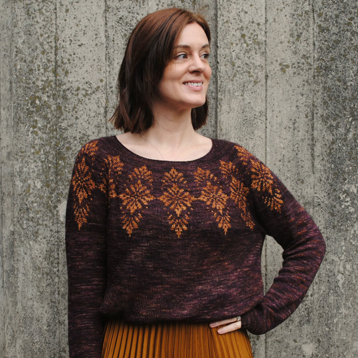A smiling woman standing in front of a grey planked background, she's wearing a beautiful colour work jumper in deep purple brown with a gold pattern and matching gold skirt