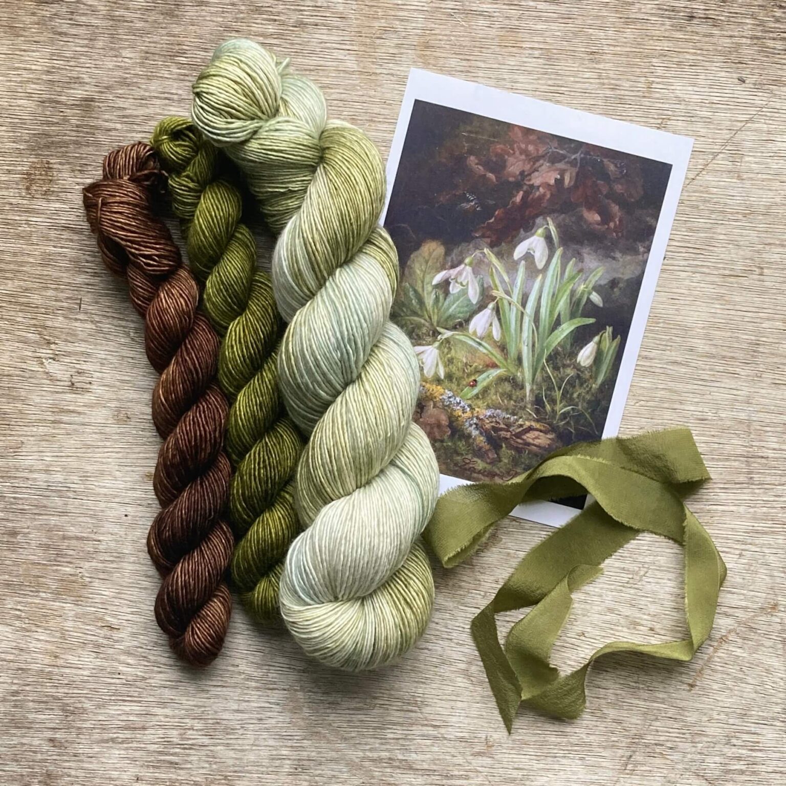 A full skein of pale green, grey and white merino and silk yarn with two minis in moss green and brown and the picture that inspired them
