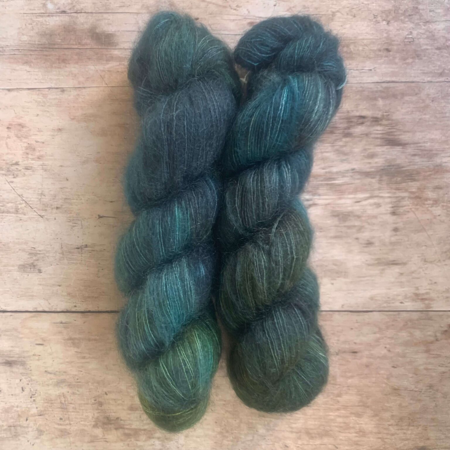 Three skeins of mid blue and green variegated mohair and silk lace weight yarn on pale wood background