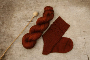 A poppy seed head, a skein of copper yarn and a hand knitted sock in the same colour yarn on a wood background surrounded by poppy seeds.