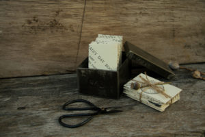 A tin box filled with hand made paper envelopes, the lid of the box leans against it and in front are a pair of scissors. To one side is a pile of envelopes tied with string and decorated with a poppy seed head.