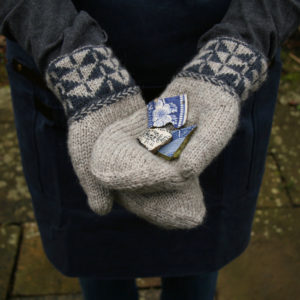 The hands of a woman crossed together with the palms upwards wearing a pair of mittens with colour work cuffs and Latvian braid and holding shards of blue and white pottery