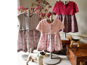 There dresses with knitted tops and floral fabric skirts surrounded by vintage toys and a suitcase