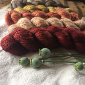 Slightly fluffy skeins of yarn in coral and red laying on a rough linen cloth with poppy seed heads in front