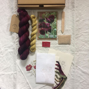 The contents of a club box laid out Dark plum yarn and a yellow mini skein, a greetings card, seed packet, handmade wooden label and an embroidery kit