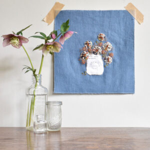 An embroidery of flowers in a jar taped to a wall with a bottle of flowers in front