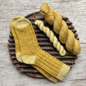 A pair of hand knit cabled socks with contrasting cuff, heels and toes and the yarn used to make them on a willow platter