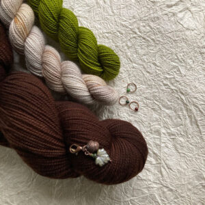 A full skein of brown wool with two minis in green and cream with a leaf stitch marker