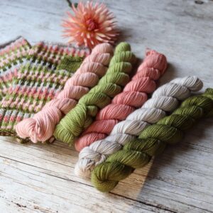 Five mini skeins in pinks and greens with a pair of fingerless gloves and a dahlia flower on a wood background
