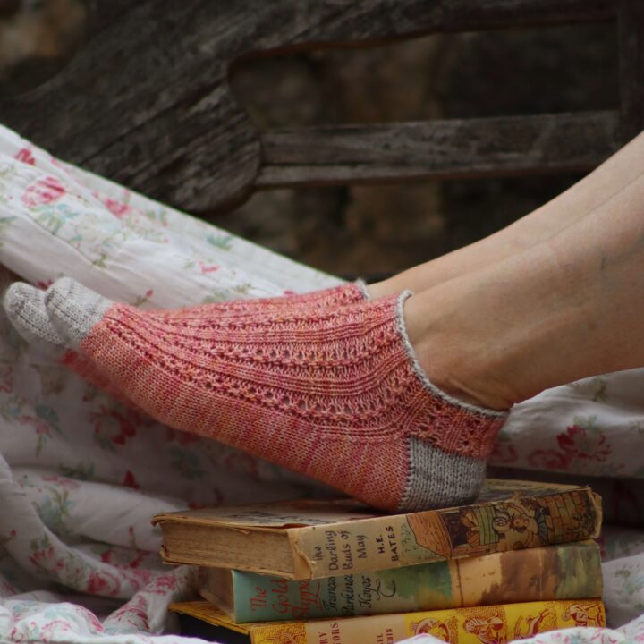 A pair of women's feet resting on a pile of books and wearing a pair of pink and stone coloured lace, ankle socks