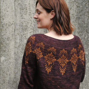 The back of a woman standing in front of a grey planked background, she's wearing a beautiful colour work jumper in deep purple brown with a gold pattern and matching gold skirt