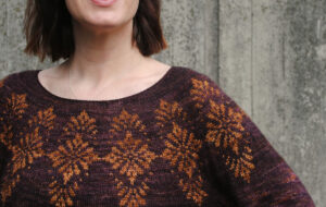 Close up of a woman standing in front of a grey planked background, she's wearing a beautiful colour work jumper in deep purple brown with a gold pattern and matching gold skirt