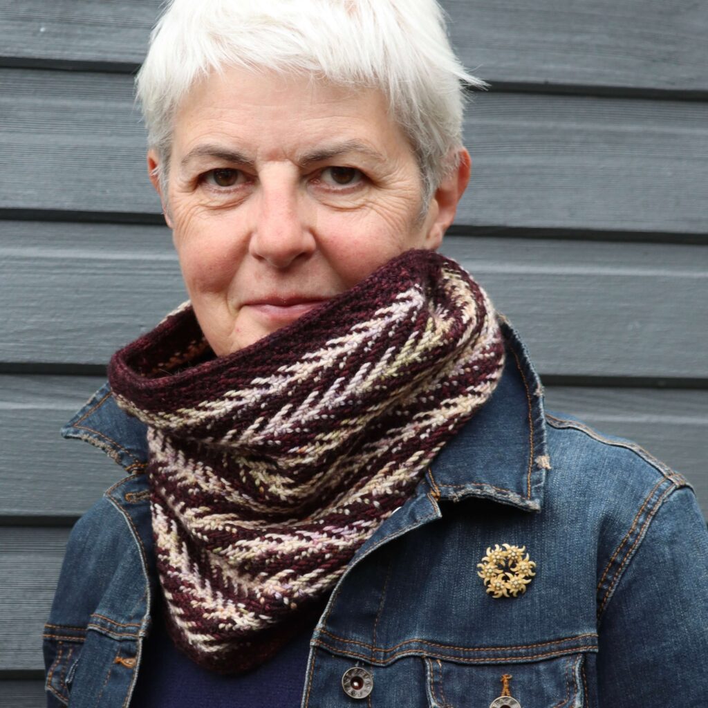A woman wearing a denim jacket and a herringbone knitted cowl looks directly at you