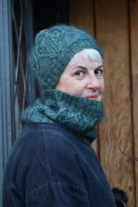 Woman in blue Heart's End bobble hat and cowl against a wooden door