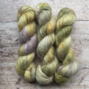 Two skeins of mohair and silk lace weight yarn in Cream, yellow, greens and lilac on pale wood background