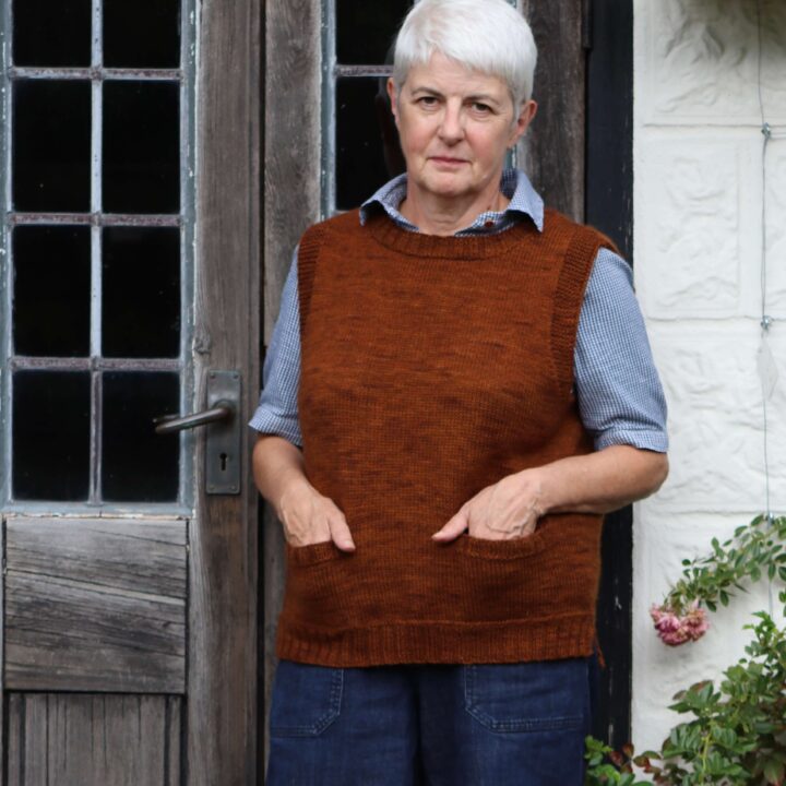 A woman standing in front of a glazed door wearing a sleeveless vest in a russet brown