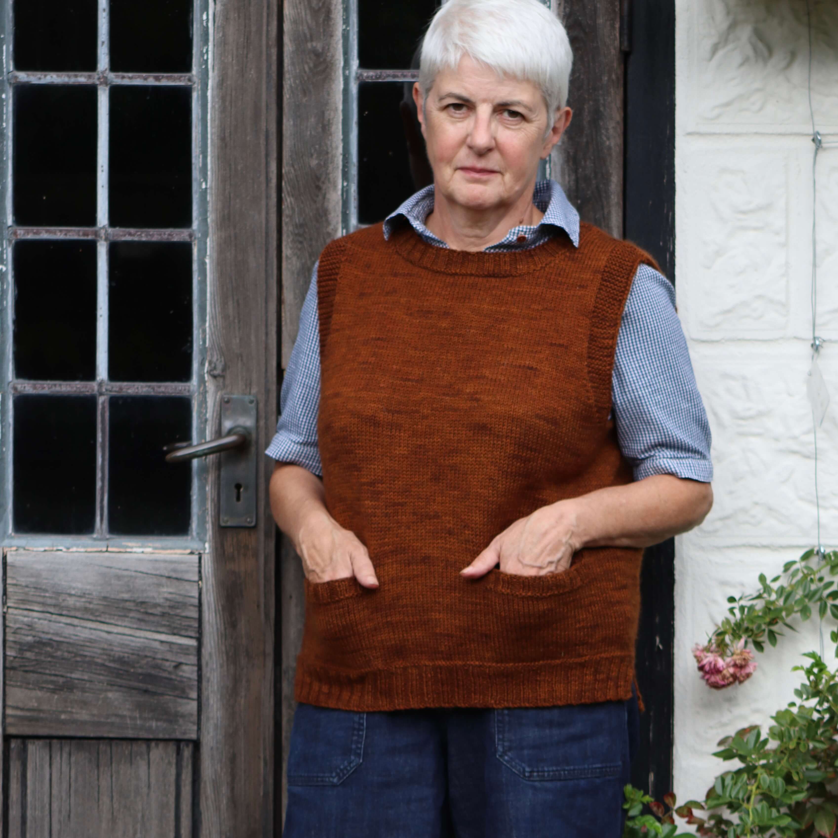 A woman standing in front of a glazed door wearing a sleeveless vest in a russet brown