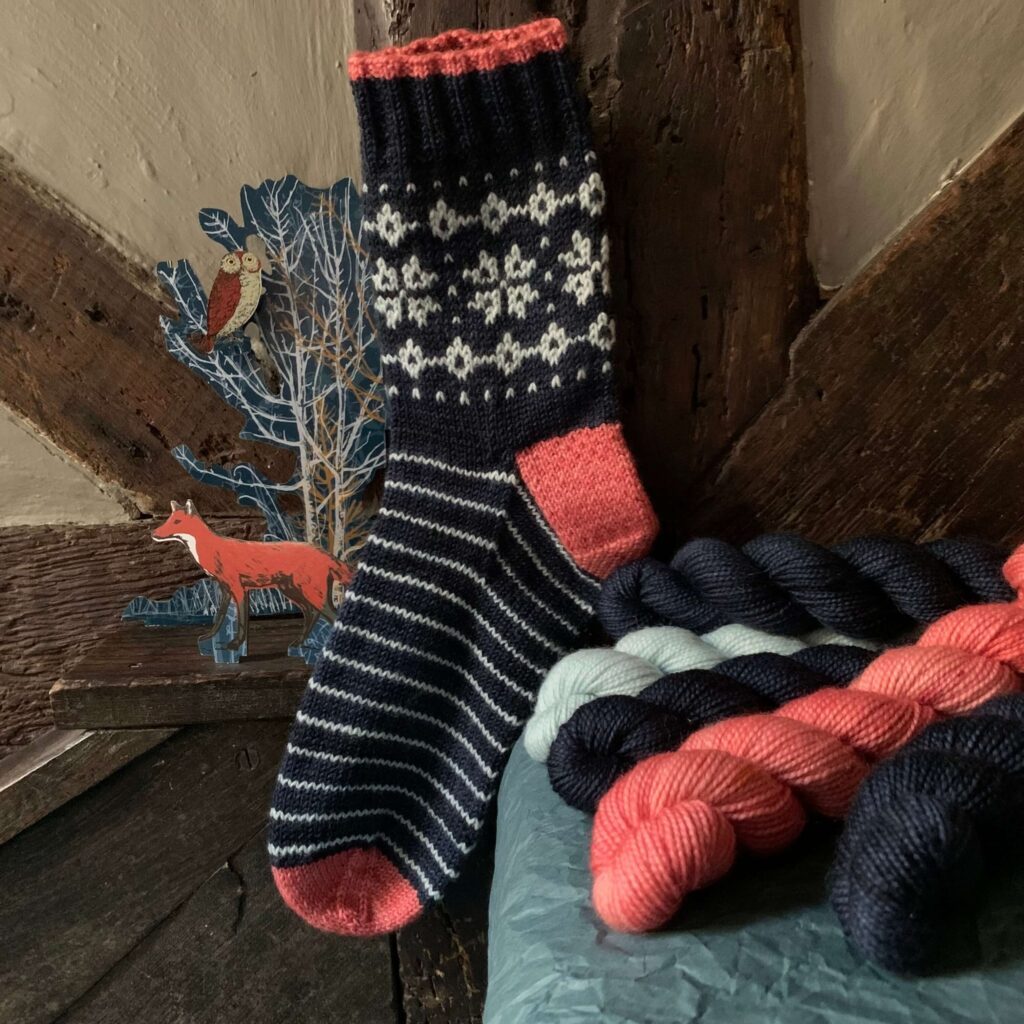 A hand knit, dark blue sock with snow crystals on it against the beams of a house with a winter cut out beside it