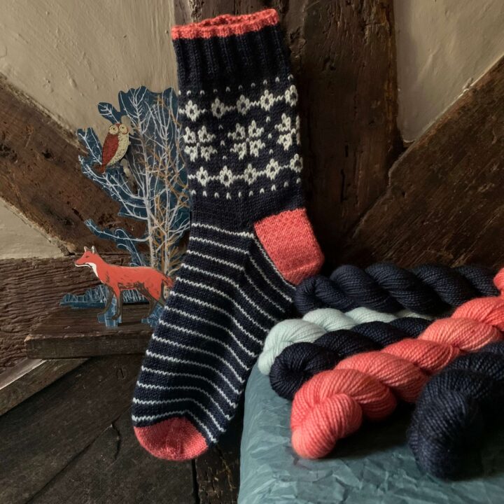 A hand knit, dark blue sock with snow crystals on it against the beams of a house with a winter cut out beside it